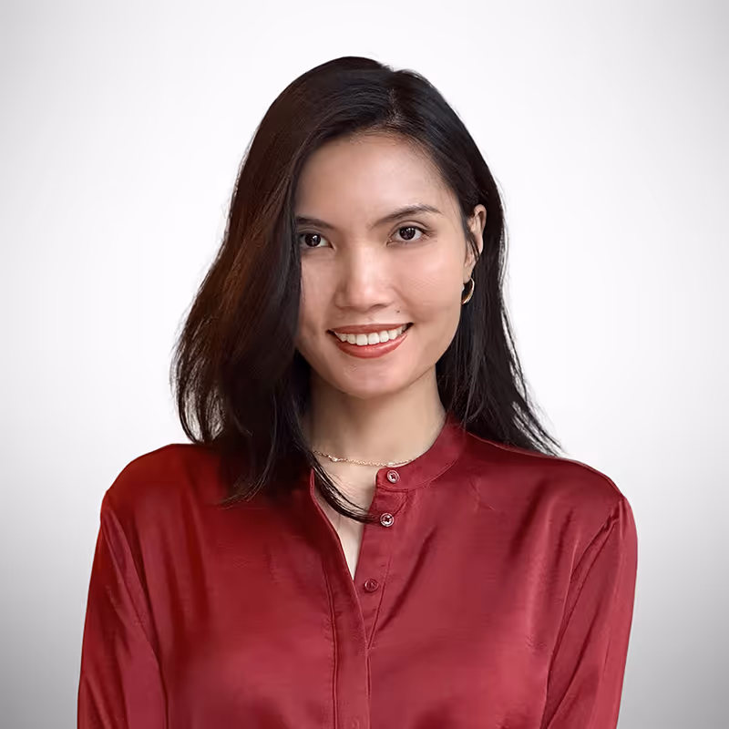 A young woman with shoulder-length dark hair smiles warmly at the camera. She is wearing a red satin blouse with a mandarin collar and gold hoop earrings. The background is a neutral gradient, and she appears confident and approachable.
