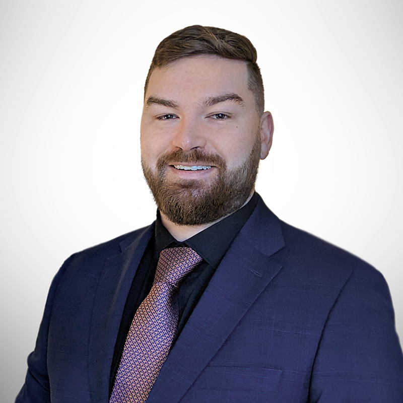 A young man with short brown hair and a well-groomed beard smiles confidently at the camera. He is wearing a navy blue suit with a black dress shirt and a patterned purple tie. His posture is professional and approachable, with a neutral gradient background.