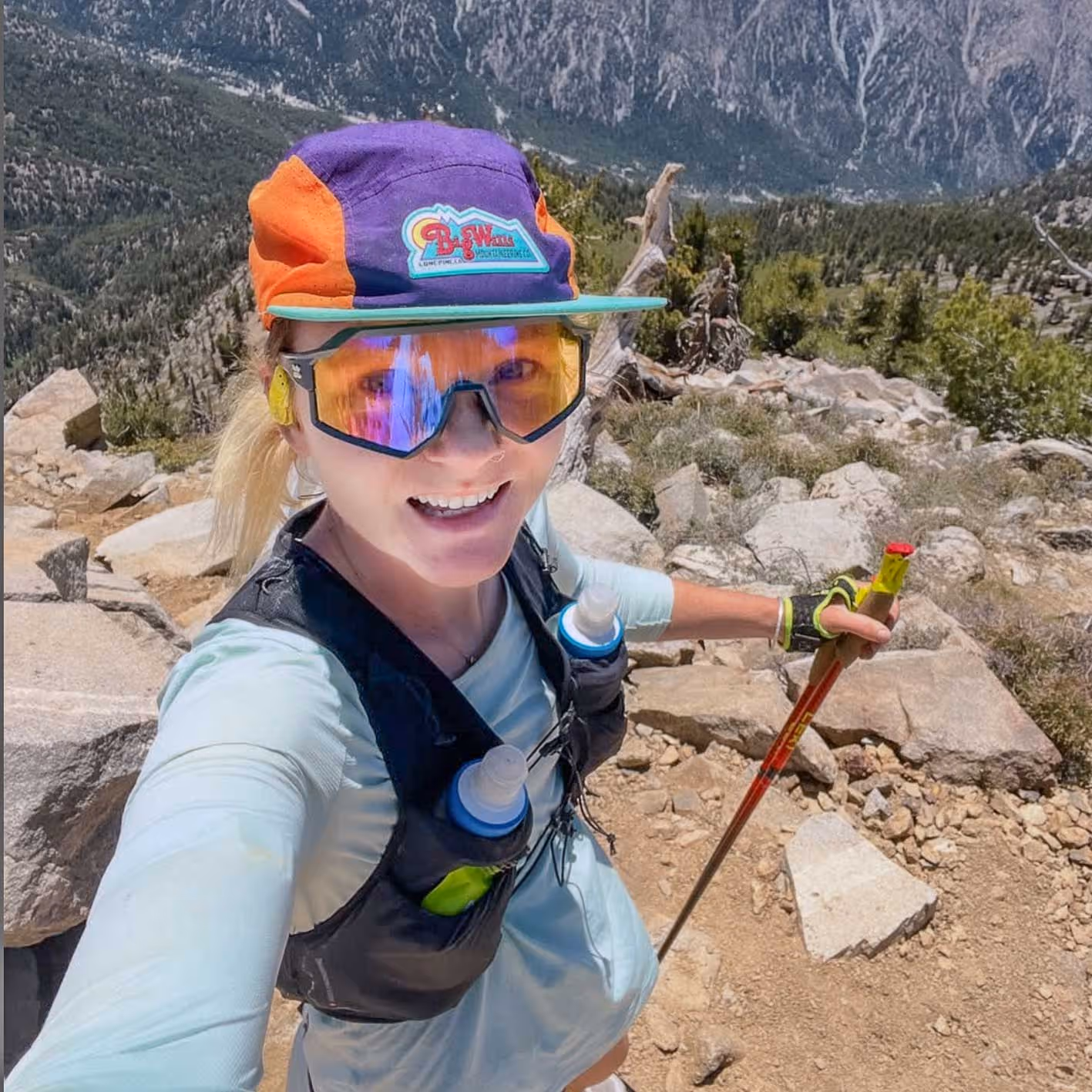 Smiling woman in sunglasses and colorful cap hiking on rocky mountain trail with trekking poles and hydration vest.