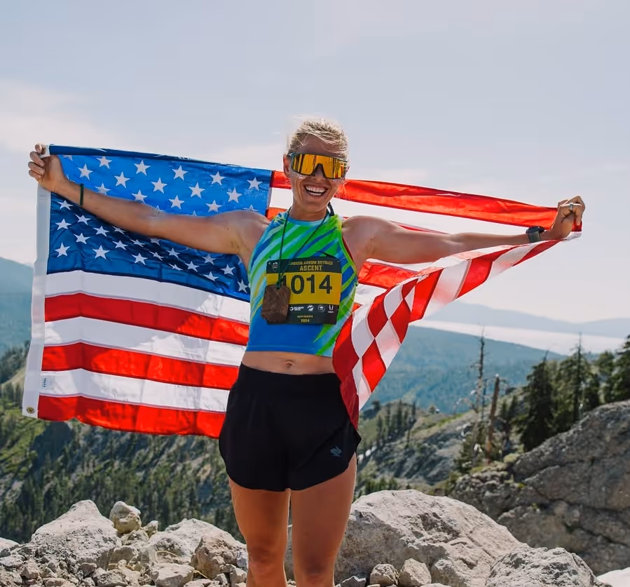 Smiling female runner wearing sunglasses and a race bib holding an American flag behind her on a rocky mountain trail.