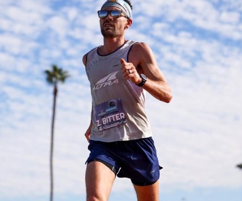 Male runner wearing sunglasses and a headband, running outdoors under a partly cloudy sky with a palm tree in the background.