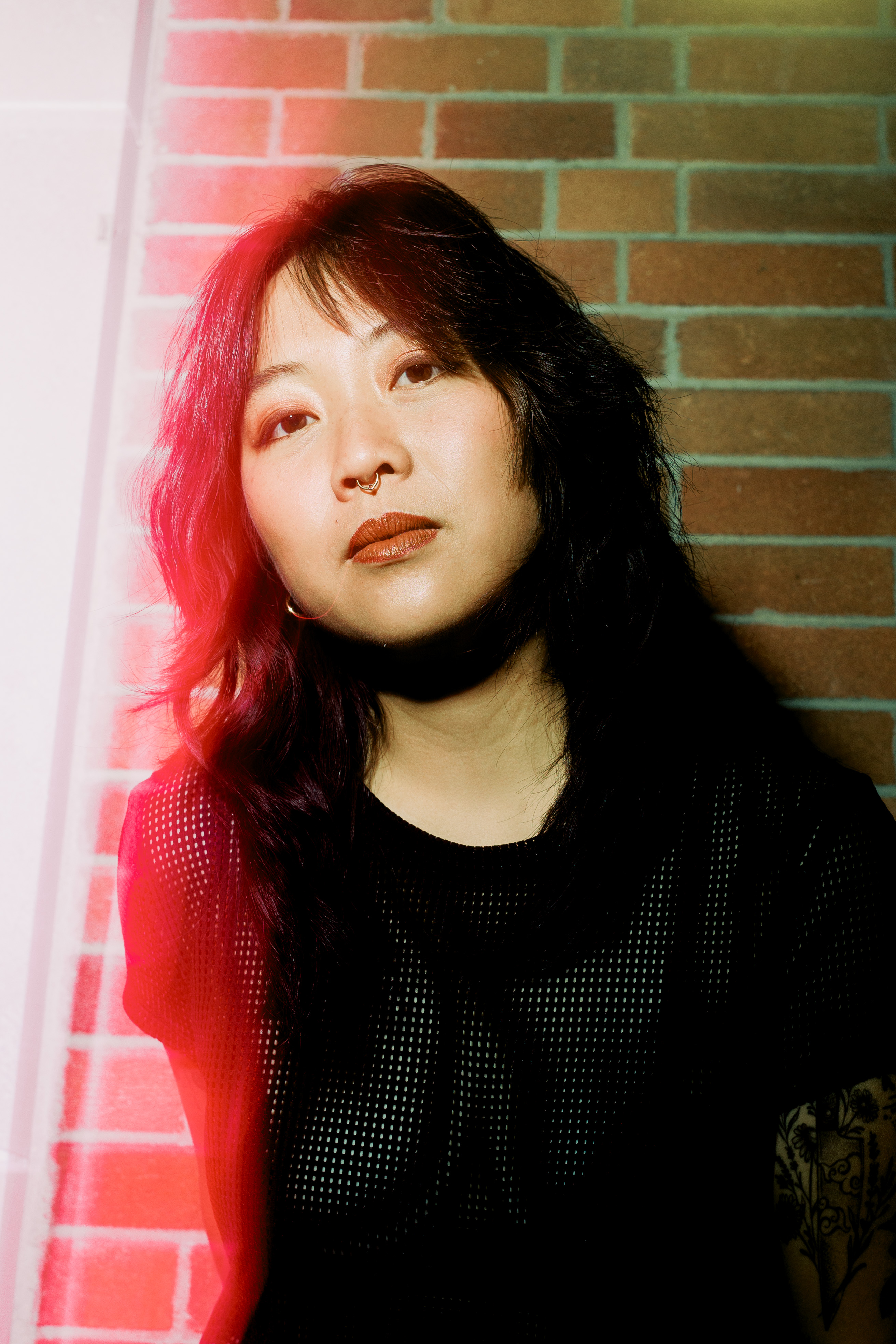 Jody, a non-binary East Asian Person with long and shaggy black hair, stands in front of a brick wall, looking into the camera. They wear a black mesh shirt, a septum ring, and brown lipstick. The left side of their body fades into a dreamy red glow. 