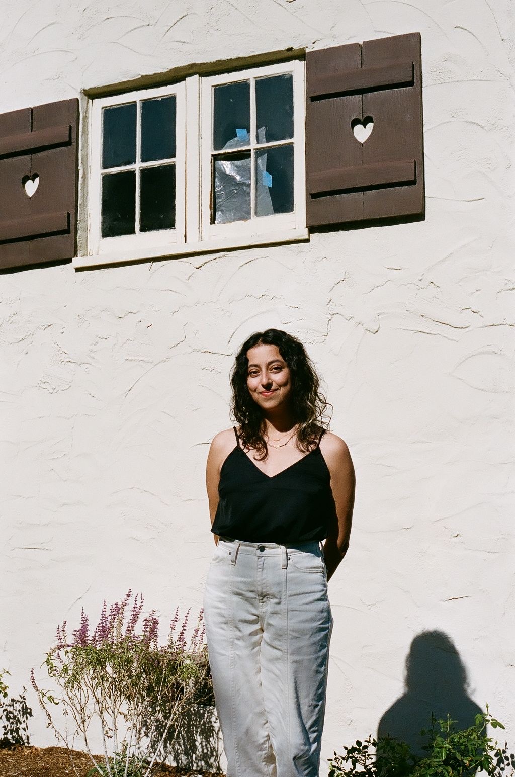 Film photo of a woman with black curly hair, standing against the white exterior of a house with her hands behind her back. Image credit: Ivanna Baranova.
