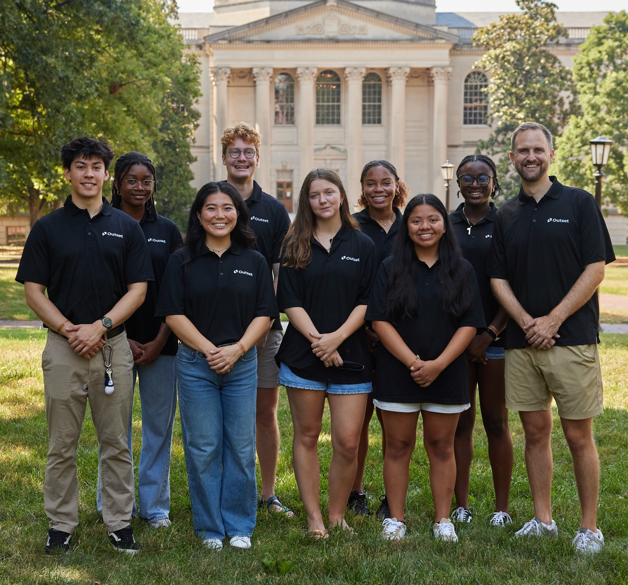 A group of eight Outset staff members stands smiling, wearing black shirts with "Outset" logos. They are outdoors on grass in front of a grand, columned building.