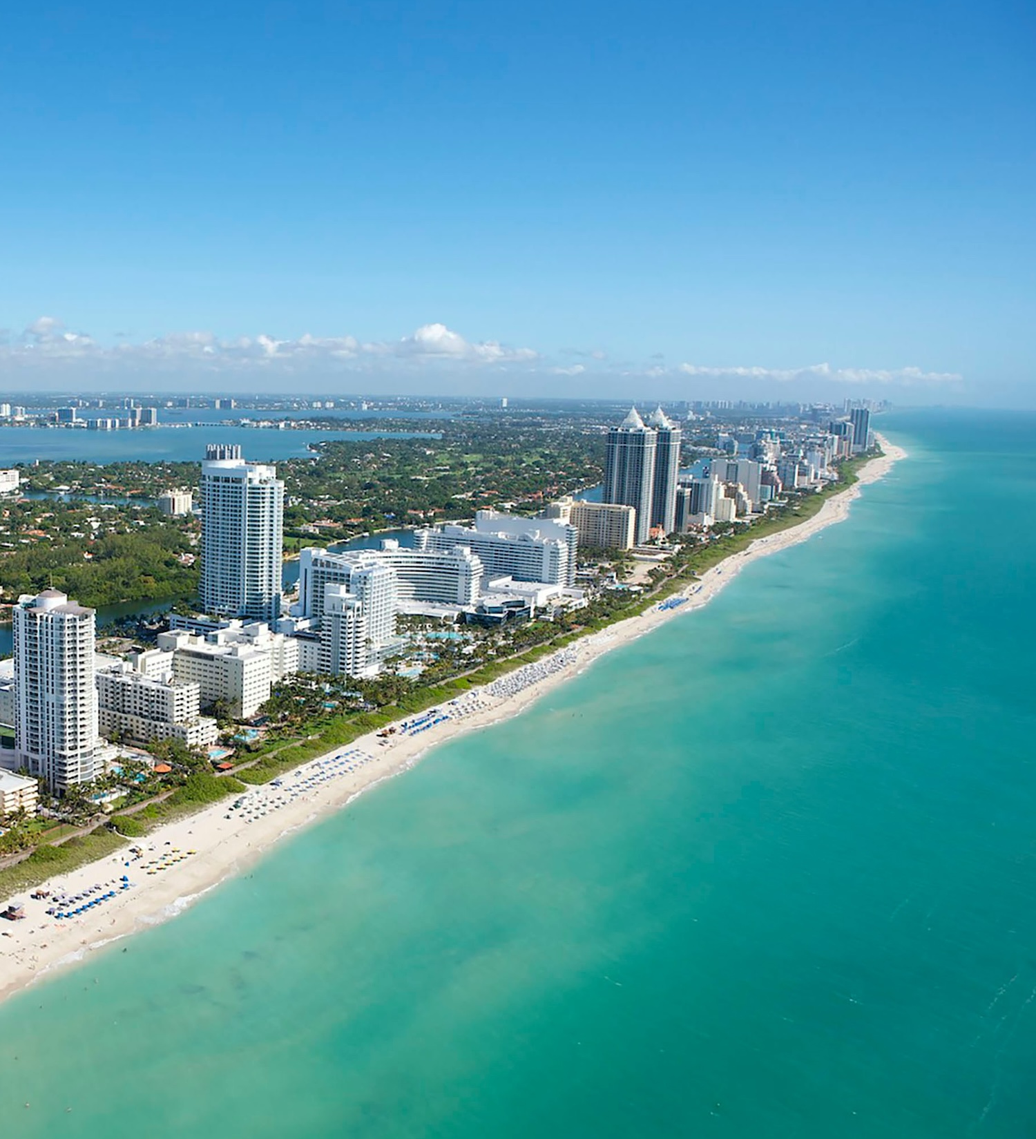 A beach lined with buildings and skyscrapers in Florida, showing lots of clear blue water and umbrellas on the sand.