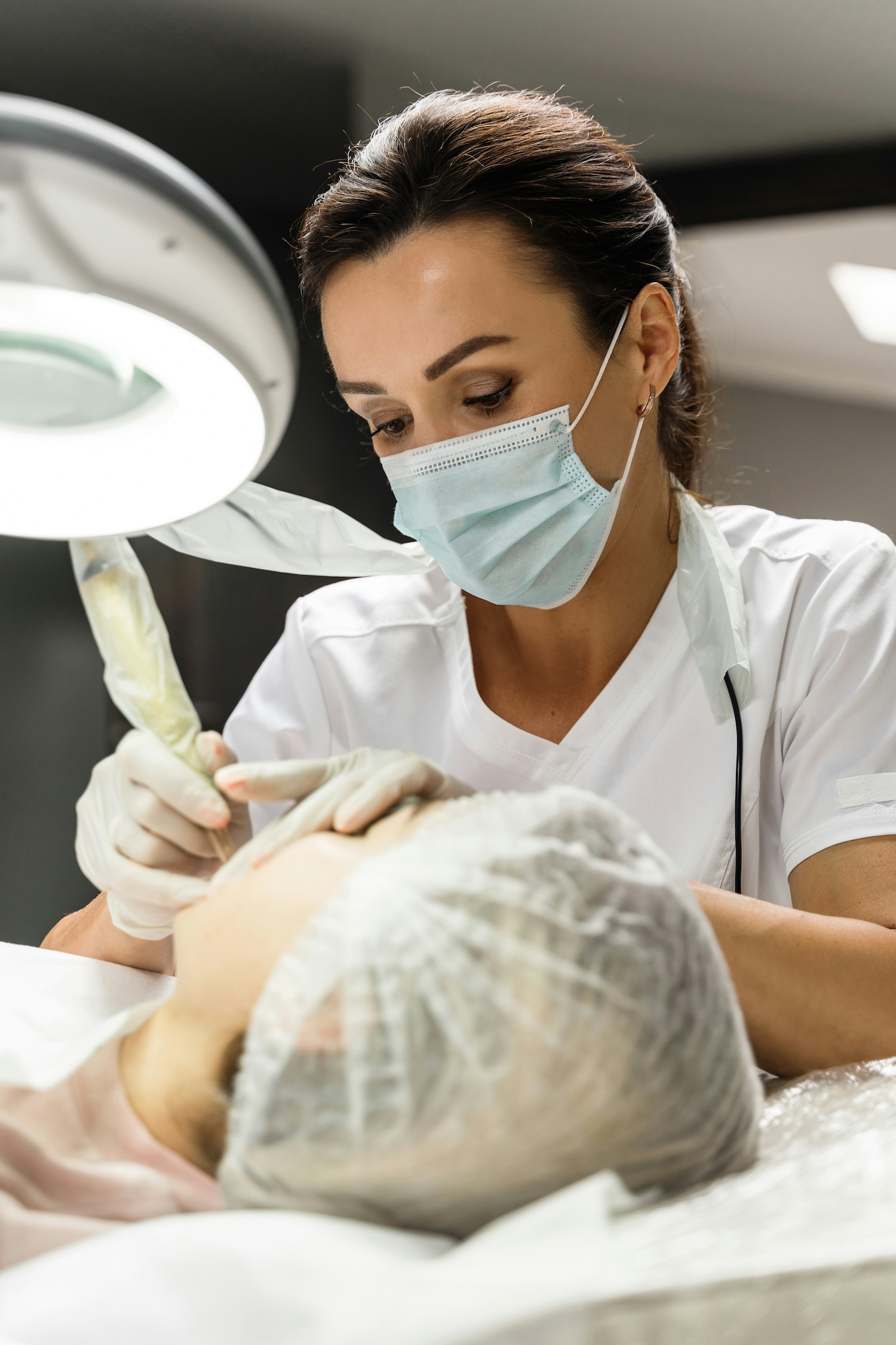 A cosmetologist wearing a mask and gloves performs a facial treatment on a client under a bright examination lamp in a clinical setting.