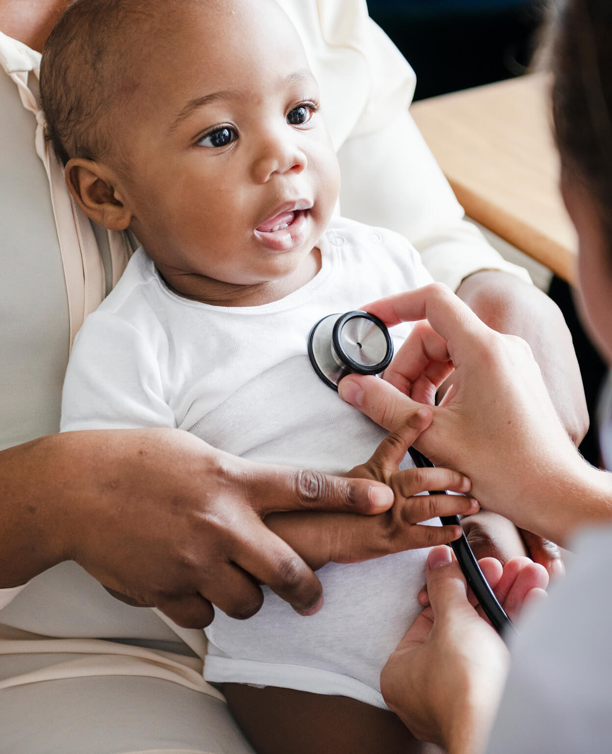 Baby sits calmly while a person uses a stethoscope on their chest. Another hand gently holds the baby's arm. They're in a clinical setting, suggesting a medical examination.