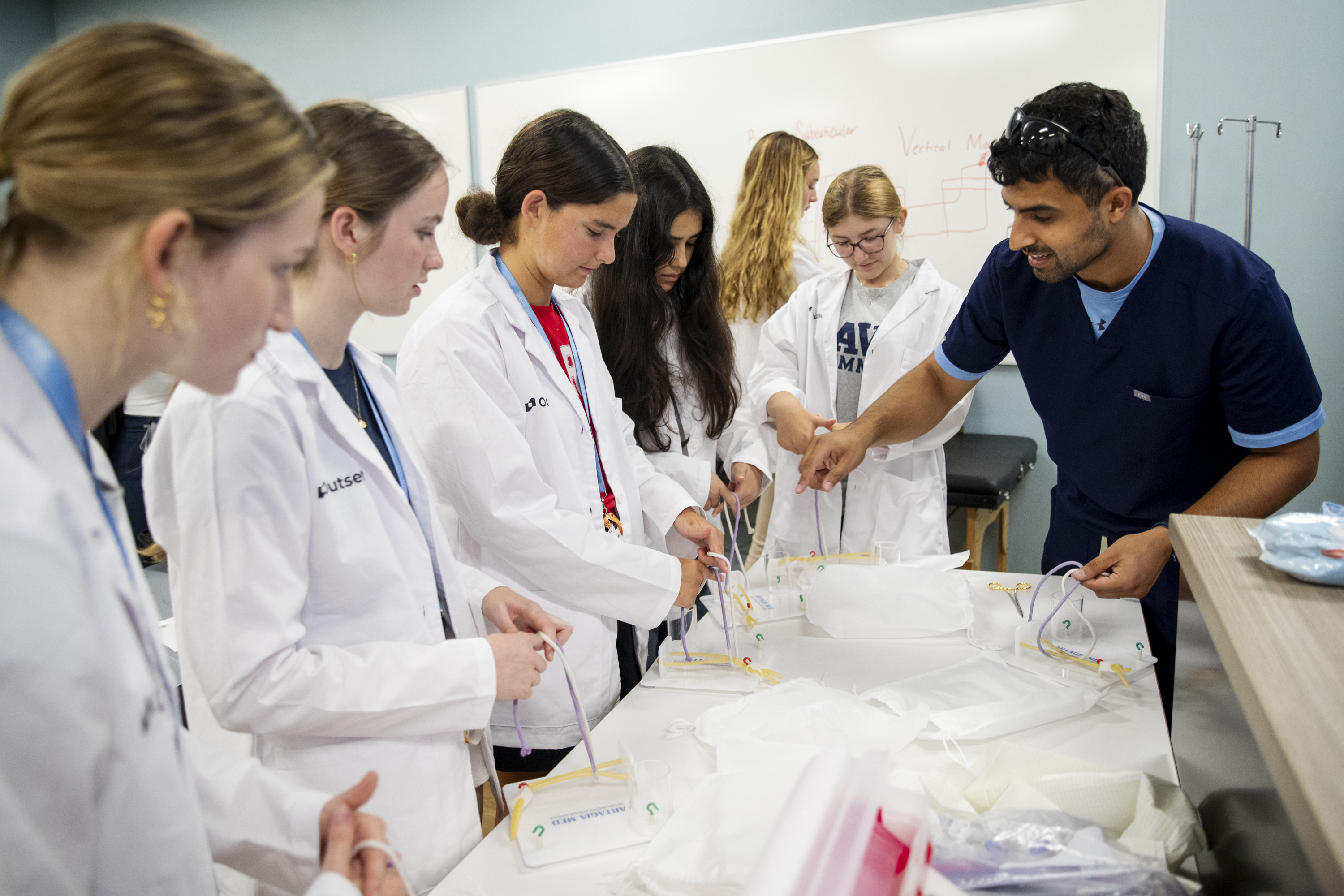 Students in lab coats practice suturing techniques with guidance from an instructor in a classroom. Whiteboards with diagrams form the backdrop.