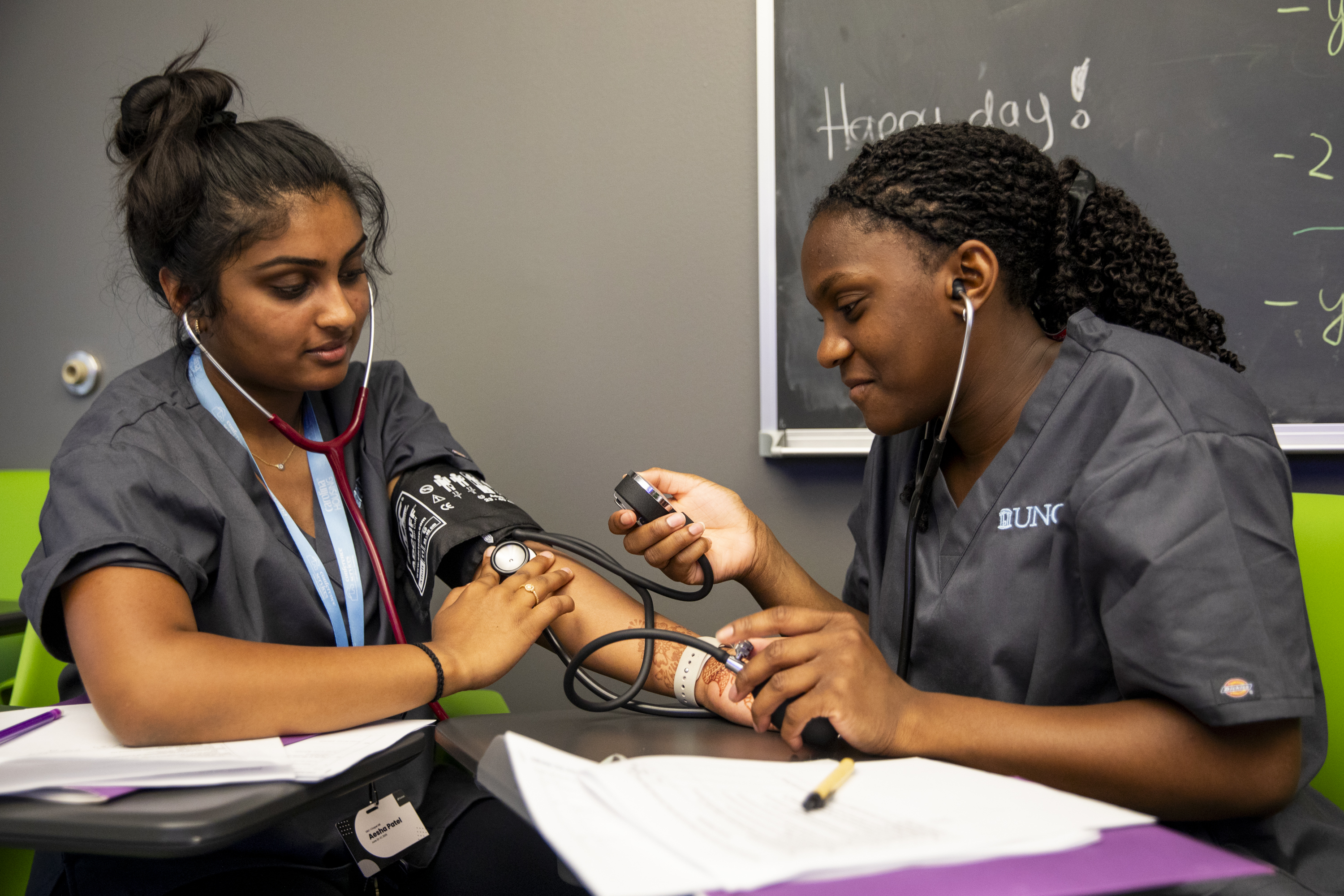 Two students in medical scrubs practice taking blood pressure; one wears a cuff, the other uses a stethoscope. They're in a classroom with "Happy day!" written on a chalkboard.