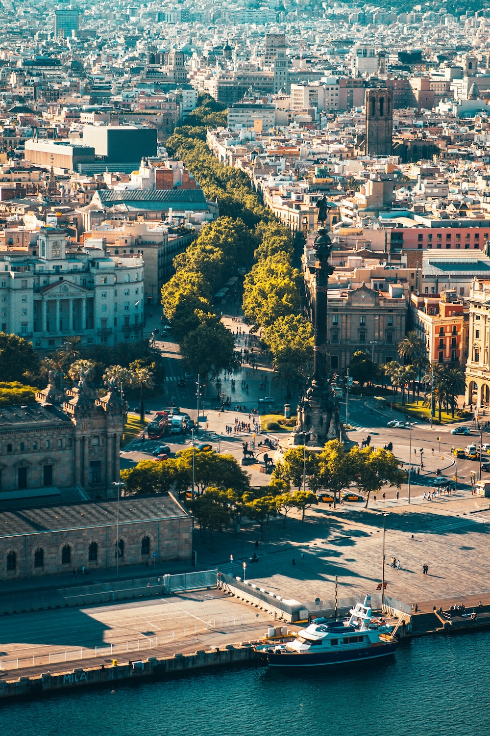 Aerial view of a cityscape shows a tall monument surrounded by trees and bustling streets. Nearby, a harbor contains a few boats. Buildings and urban sprawl fill the background.