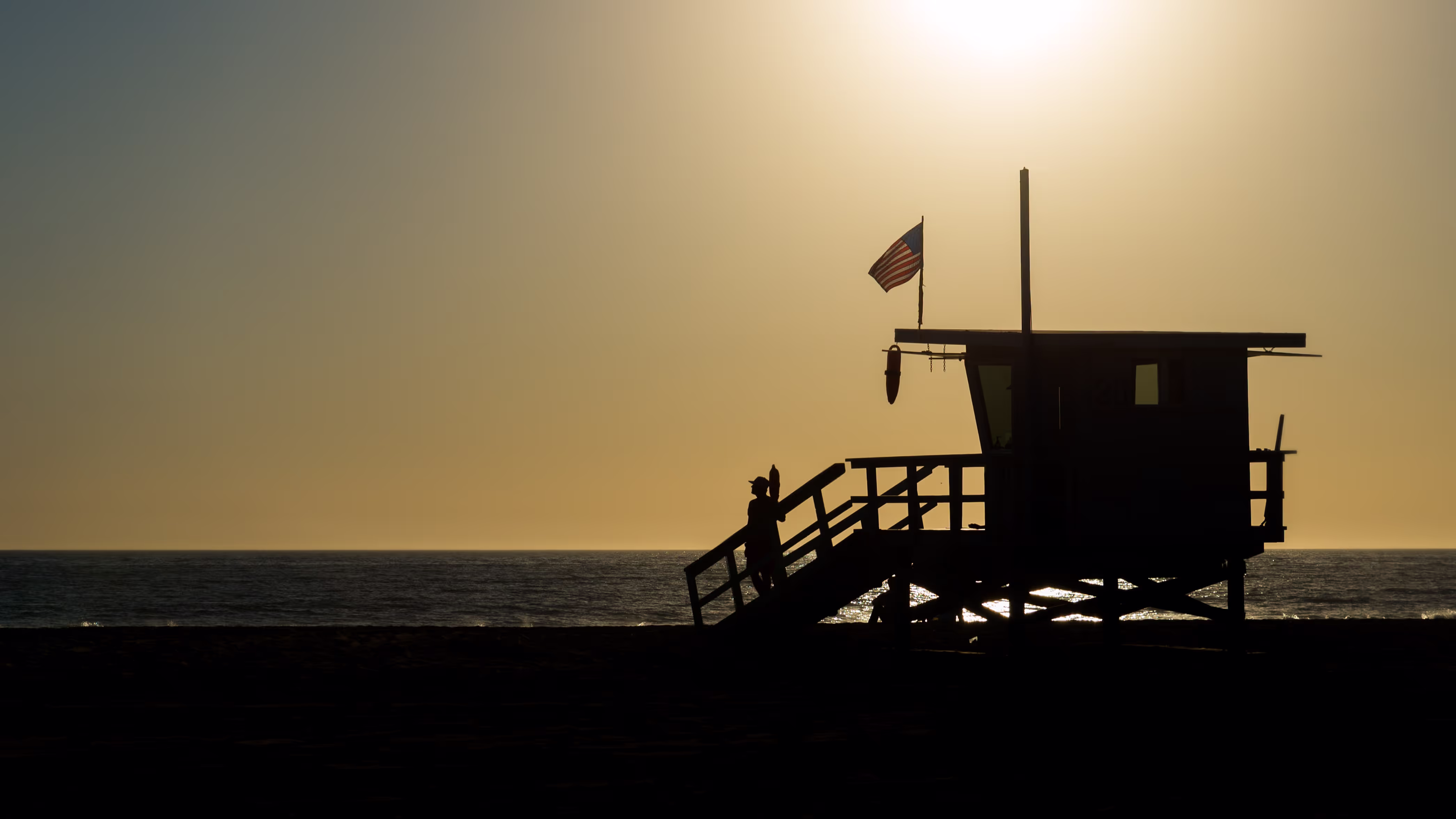 Silhouette of lifeguard tower with American flag at sunset by ocean