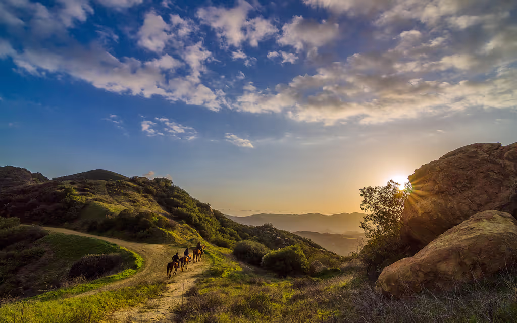 Horseback riders on trail at sunset with dramatic mountain and cloud landscape