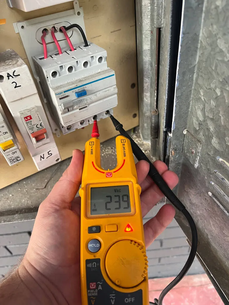 Hand holding a yellow digital multimeter measuring voltage on an electrical circuit breaker panel