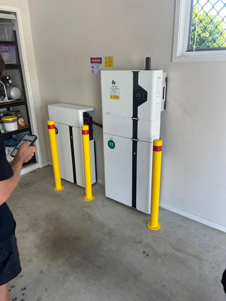 Two white electrical battery storage units mounted on a wall with yellow safety bollards in front and a person holding a phone nearby