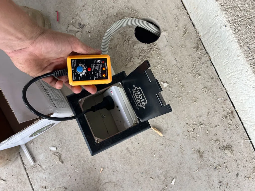 Hand holding an electrical tester connected to a power outlet box embedded in a concrete floor