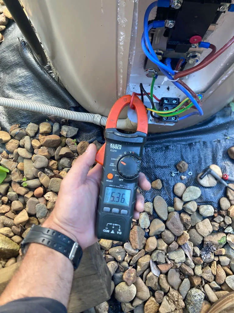 Hand holding a Klein Tools clamp meter measuring electrical current near wiring and pebbles on the ground