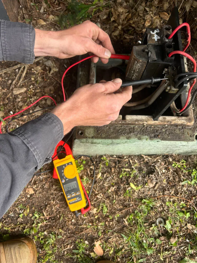 Hands holding red and black electrical test leads connected to an outdoor electrical component with a yellow multimeter on the ground