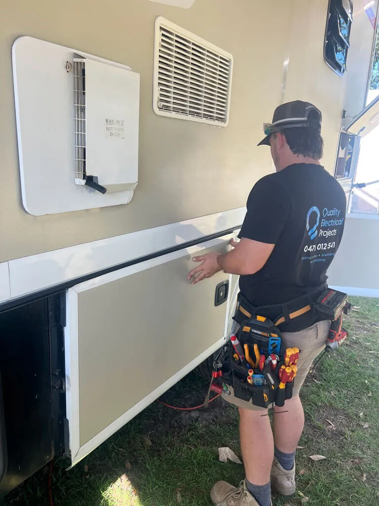 Electrician wearing a Quality Electrical Projects shirt working on the side panel of a caravan with tools in a belt