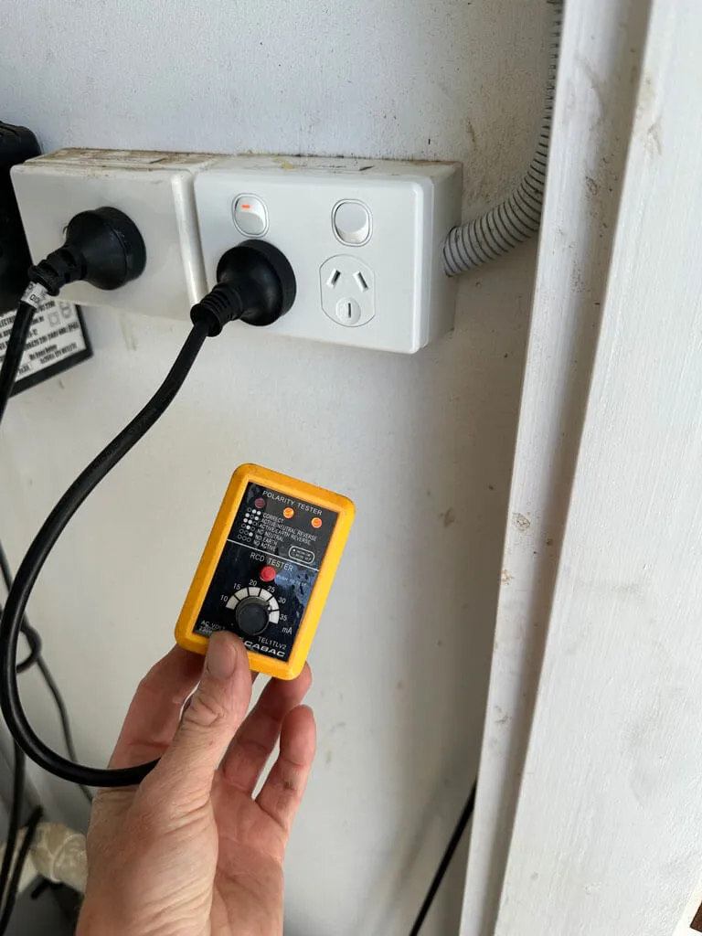 Hand holding a yellow electrical tester near a double power outlet with two plugs inserted