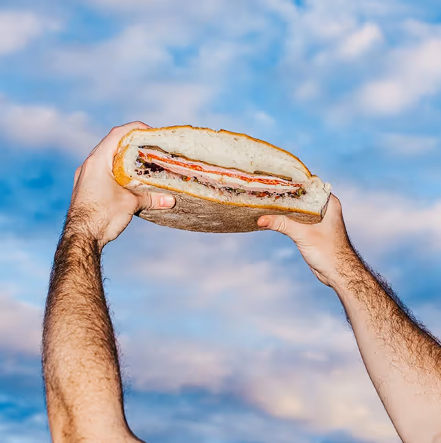 Person holding deli sandwich on similar blue cloudy sky aesthetic background like other ads