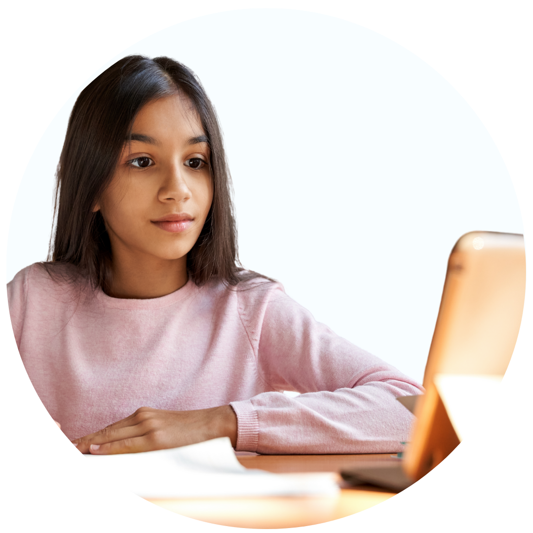 Young girl in a pink sweater attentively looking at a tablet screen while sitting at a desk with papers.