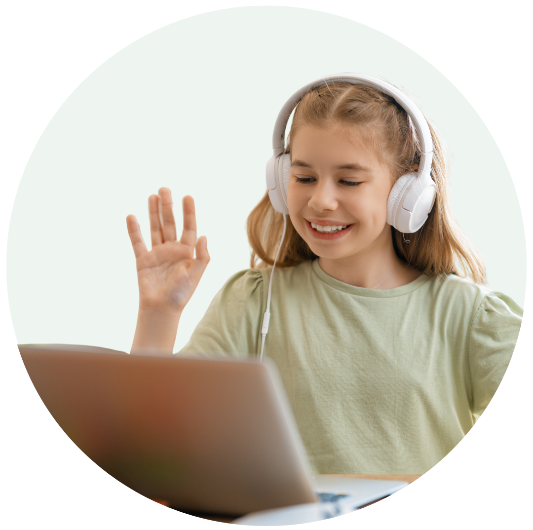 Young girl wearing white headphones smiling and waving at a laptop screen during a video call.