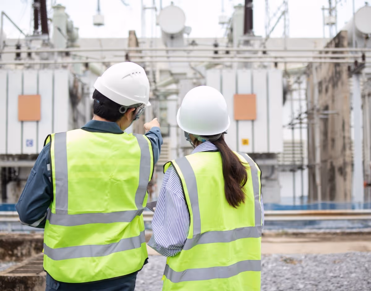 Two workers in safety vests and helmets inspect electrical substation equipment