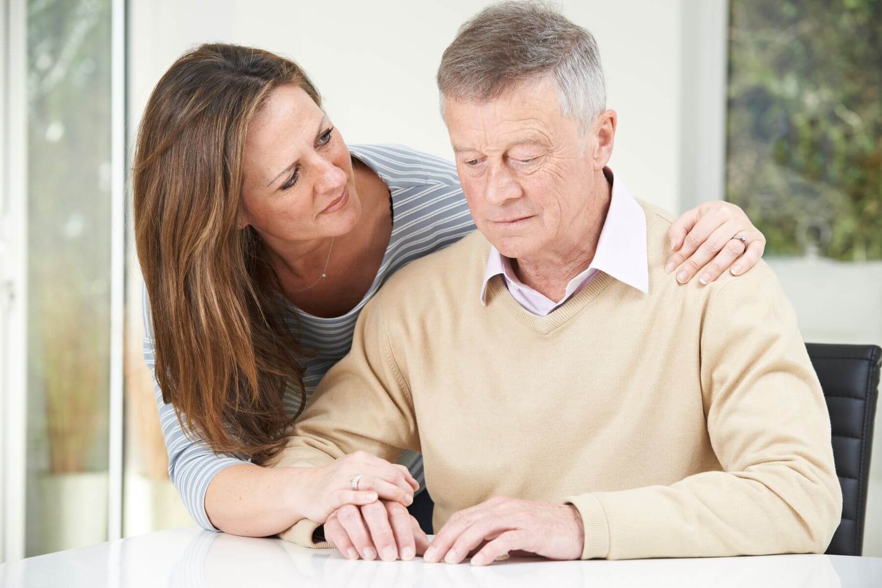 An elderly woman hugging an elderly man who is sat at a table