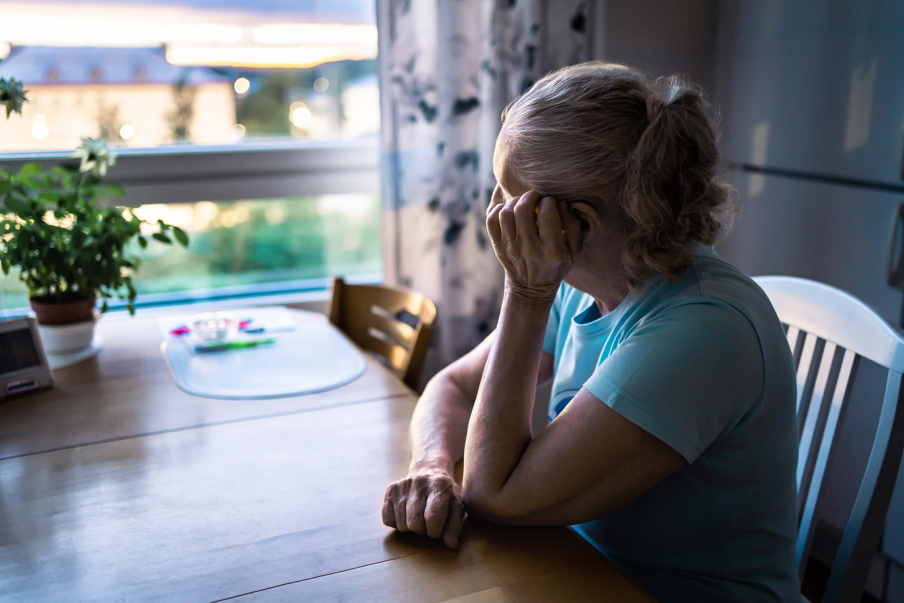 An elderly woman sitting at a dining table, resting her head on her hand and staring out of a window