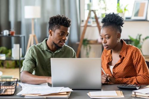 Two people reviewing paperwork together at a laptop in a home office setting.