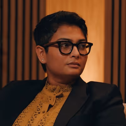 Person with short hair wearing black glasses, a mustard yellow lace top, and a dark blazer looking to the side against a warm wooden background.