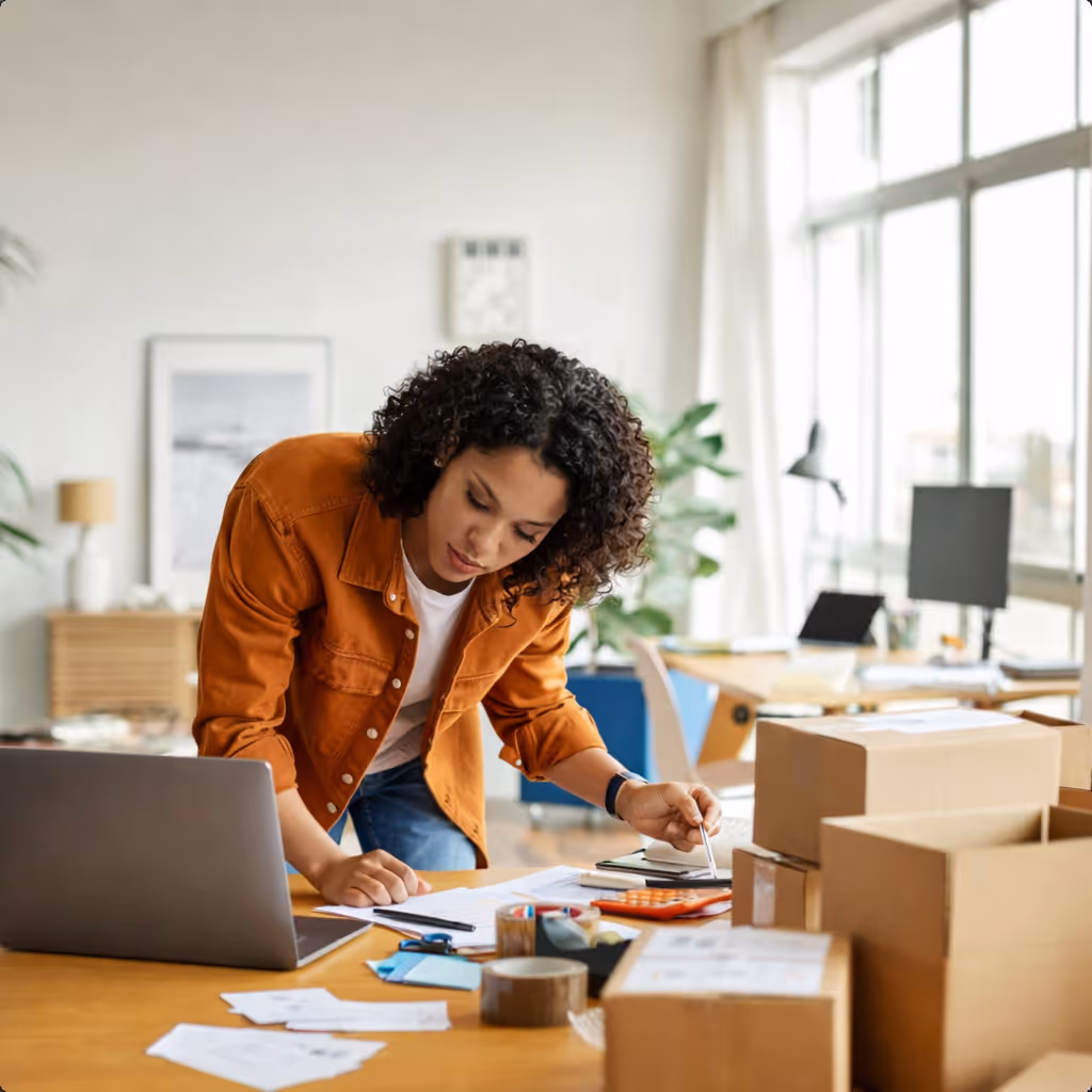 Woman in orange jacket working at a desk with paperwork, laptop, and cardboard boxes in a bright office.