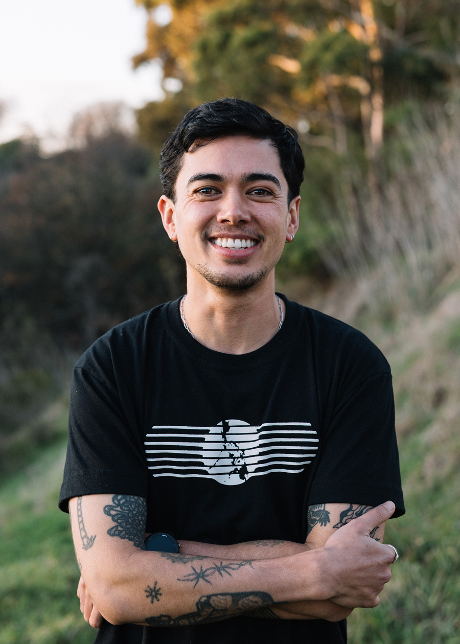 Portrait of Rio, a light brown person smiling. They have dark short hair and their arms are crossed with tattoos and a black t-shirt.
