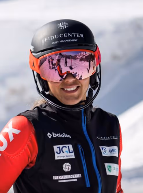Smiling skier wearing a black Fiducenter helmet and reflective goggles, dressed in black and red ski gear with sponsor logos.