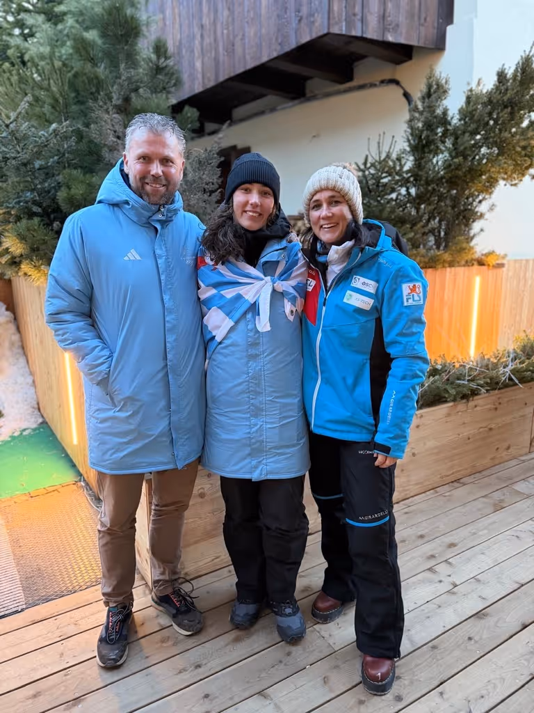 Three people wearing winter jackets and hats standing on a wooden deck with trees and fencing in the background.