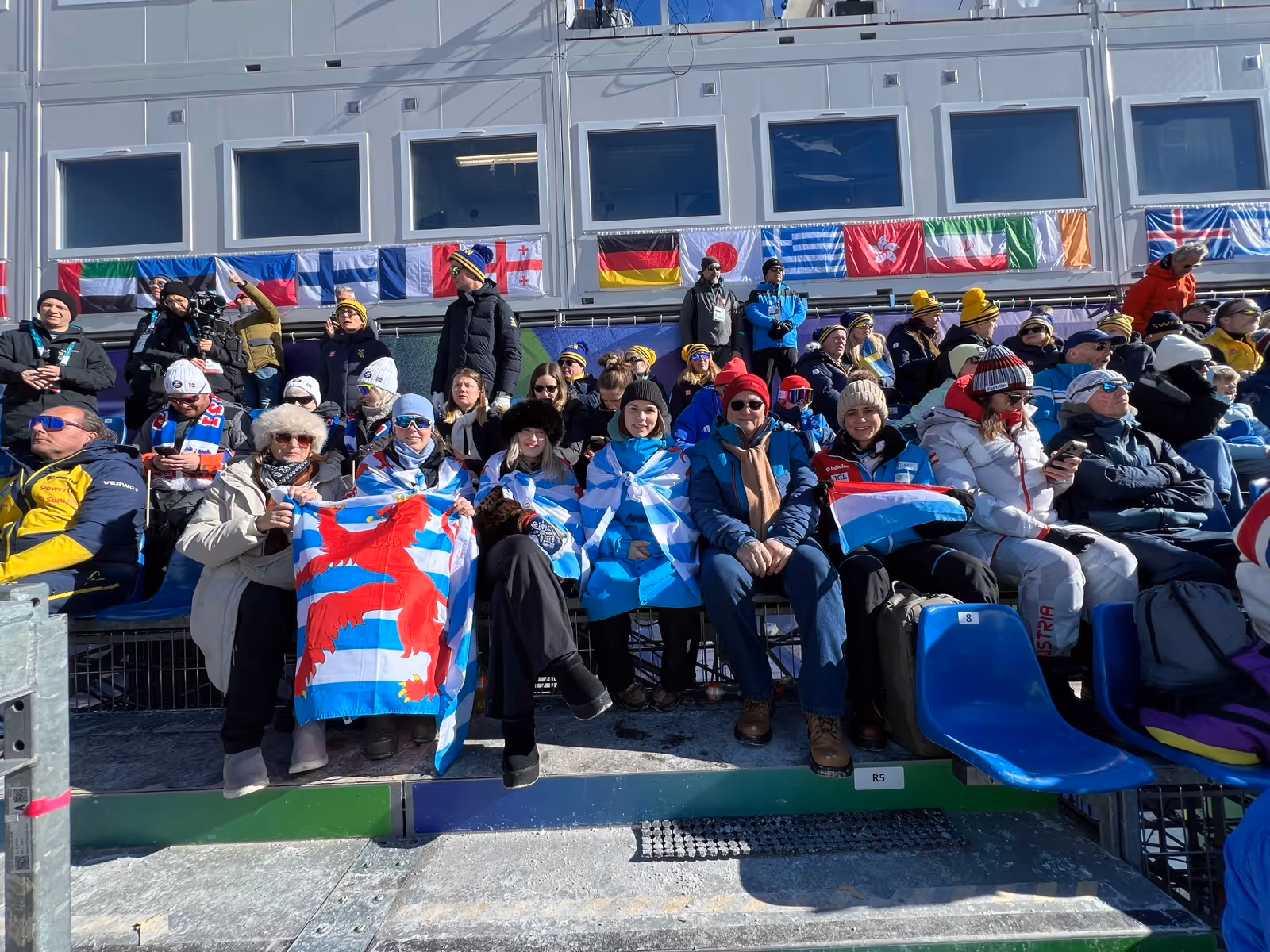 Spectators bundled in winter clothes sitting in stadium seating under flags from various countries on a sunny day.