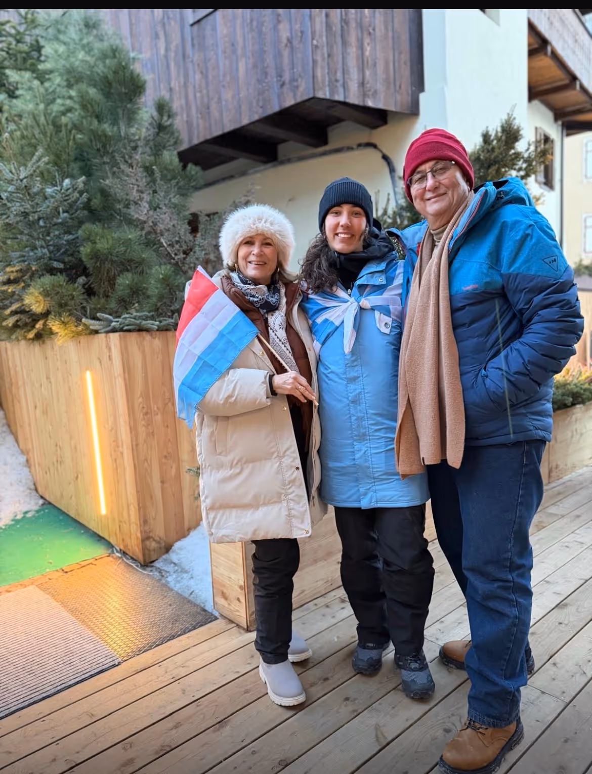 Three people wearing winter coats and hats stand on a wooden deck, smiling; one holds a small blue, white, and red flag.