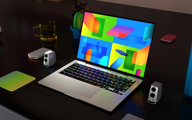 Open laptop on a dark desk displaying a colorful abstract screensaver with geometric shapes, surrounded by two small speakers, a mousepad, a cactus plant in a blue pot, a glass, and a closed book.