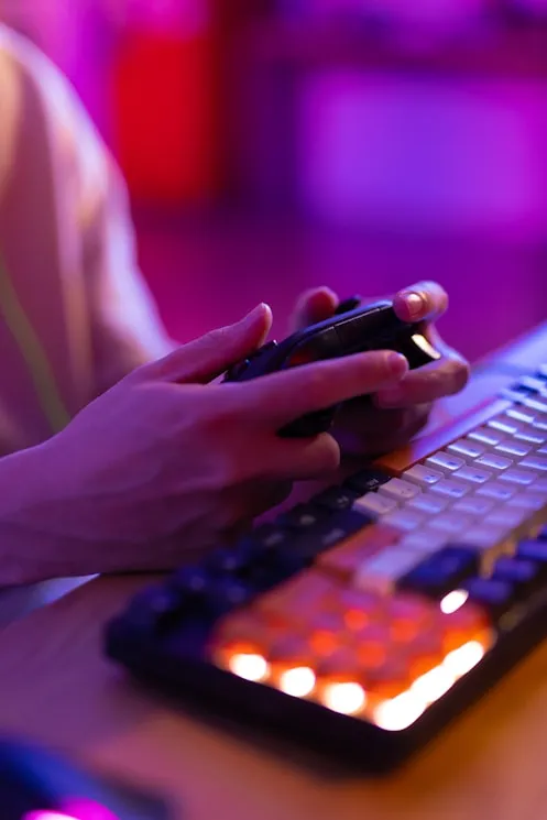 Person holding a game controller near a backlit computer keyboard with colorful lighting in a dimly lit setting.