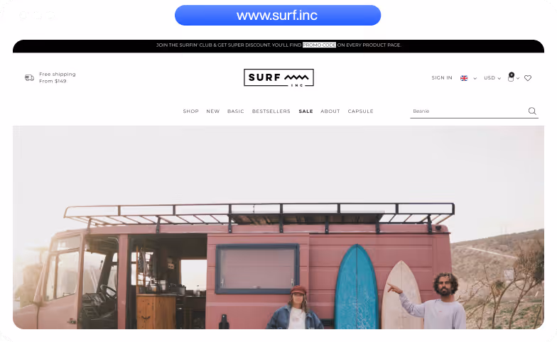 Two people standing in front of a pink camper van with two surfboards, one blue and one beige, leaning against it in an outdoor setting.