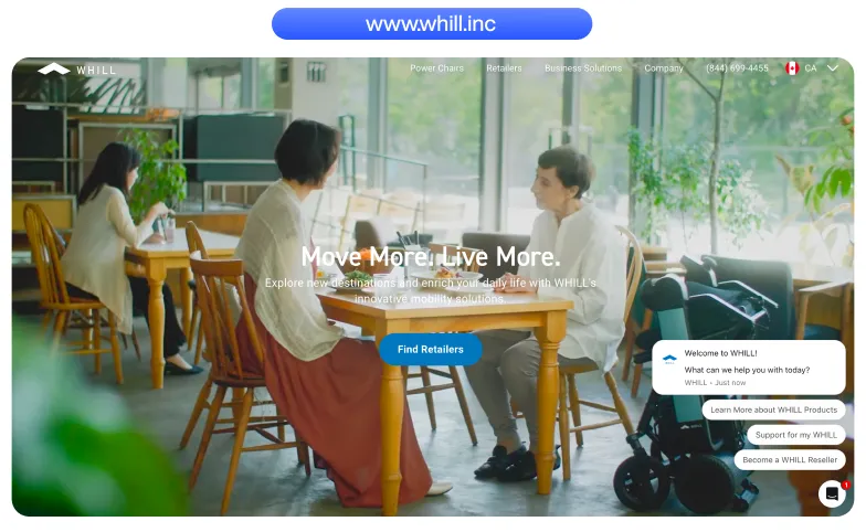 Two people sitting at a wooden table in a bright cafe, with a WHILL mobility device beside one person.