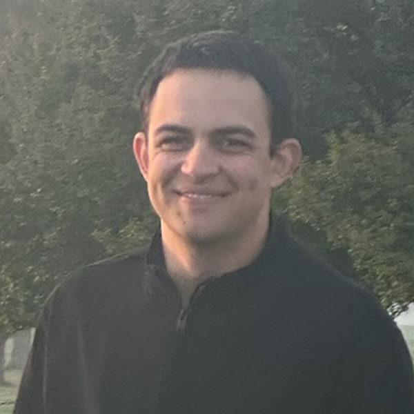 Man with short dark hair smiling outdoors with trees in the background.