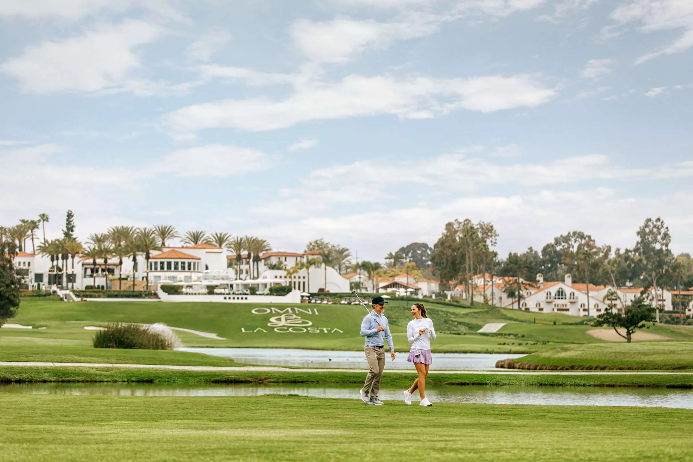 Man and woman walking on a golf course near a water hazard with Omni La Costa resort buildings behind.