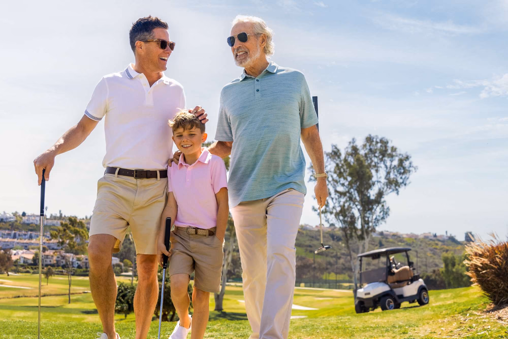 Three generations of men walking on a golf course holding golf clubs and smiling.
