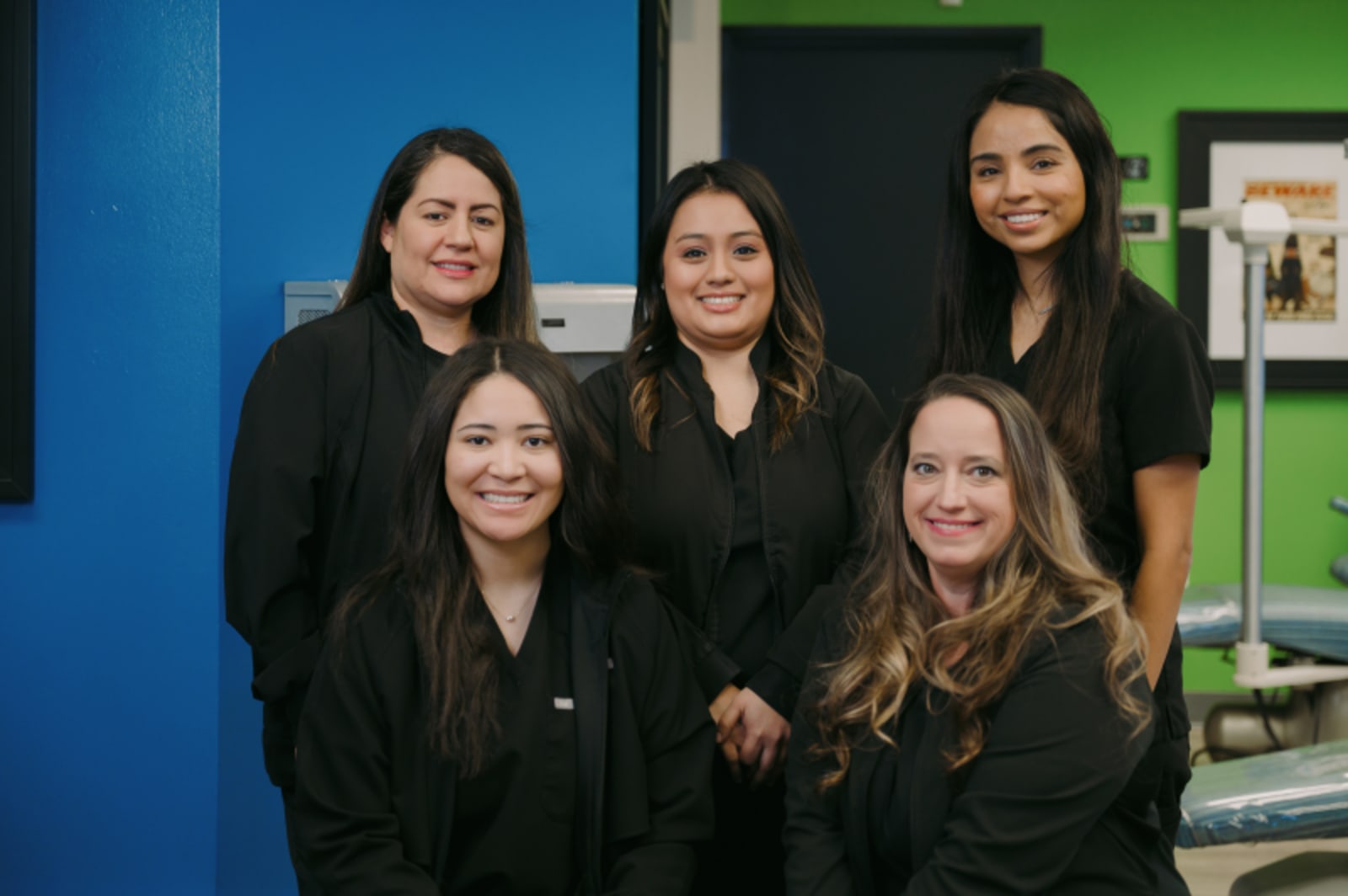 Five female dental team members smiling together in an office setting with blue and green walls.