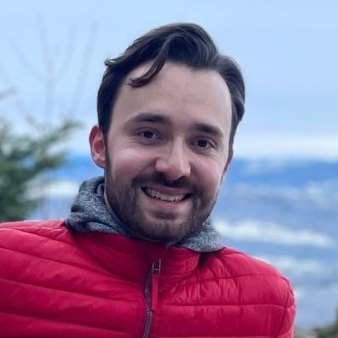Smiling man with dark hair and beard wearing a red jacket outdoors with blurred landscape background.