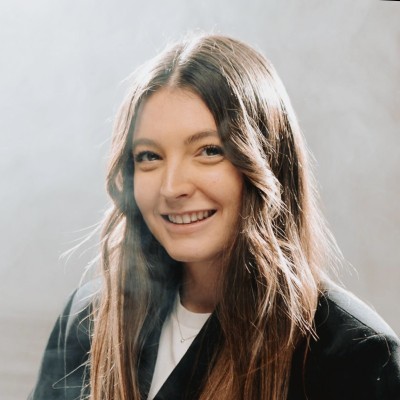 Young woman with long brown hair smiling, wearing a black jacket and white top against light background.