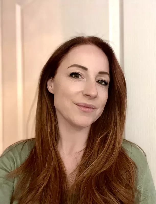 Woman with long red hair and a green top smiling gently against a light background.