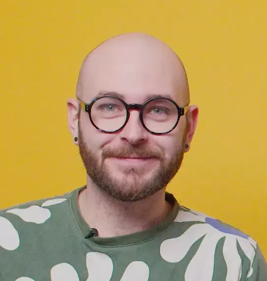 Smiling bald man with a beard and round glasses wearing a patterned green shirt against a yellow background.