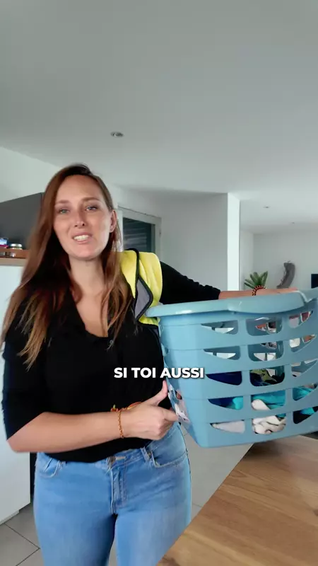 Woman in black shirt and jeans holding a blue laundry basket with clothes indoors.